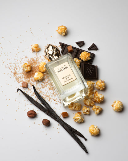 A bottle of Brownie Pop Extrait de Parfum by Fragrapedia Haus rests among popcorn, caramel, chocolate, vanilla beans, hazelnut, and brown sugar—capturing the deliciously decadent essence of a brownie-inspired gourmet scent.