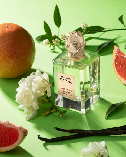 A Maison Mataha Printemps Blanc Extrait de Parfum bottle rests on a green surface, surrounded by white flowers, grapefruit halves, vanilla beans, and green leaves—evoking the fragrance’s floral notes with jasmine sambac hints.