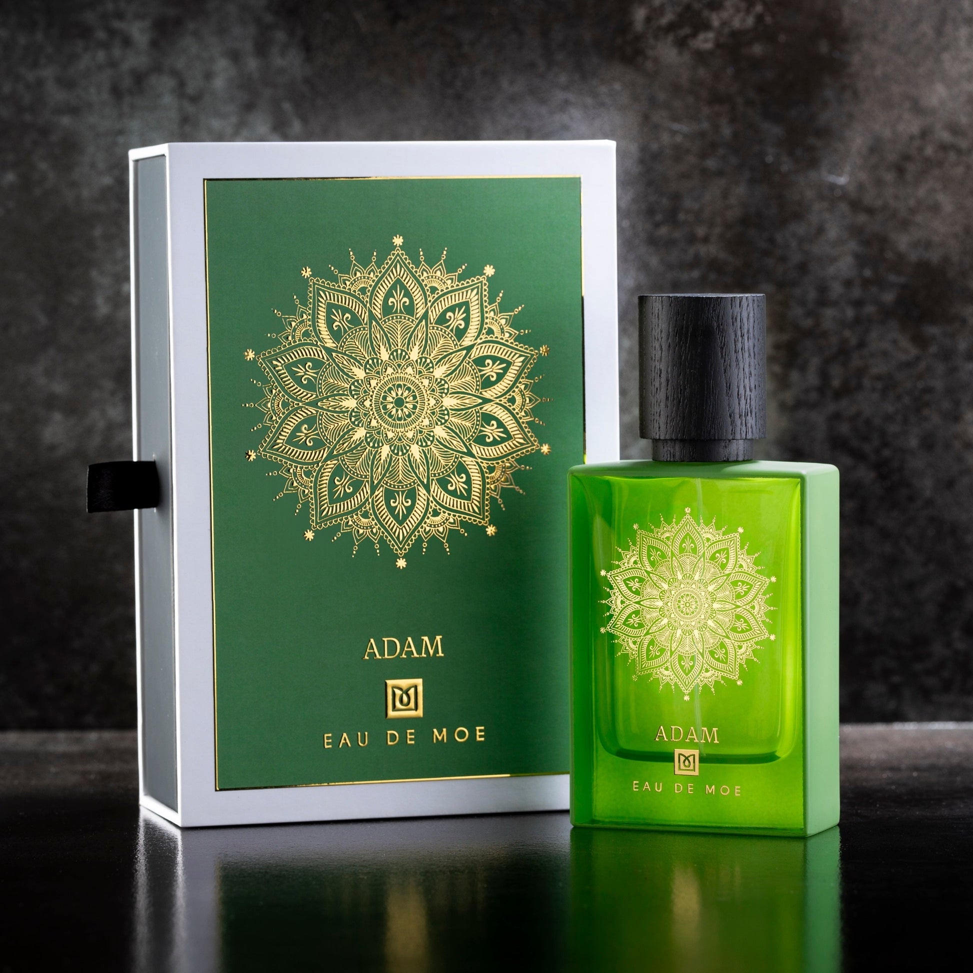 A green ADAM Eau de Parfum bottle by Eau De Moe stands beside a matching green and gold mandala-patterned box. This men’s fragrance features Italian bergamot notes and is showcased on a dark surface.
