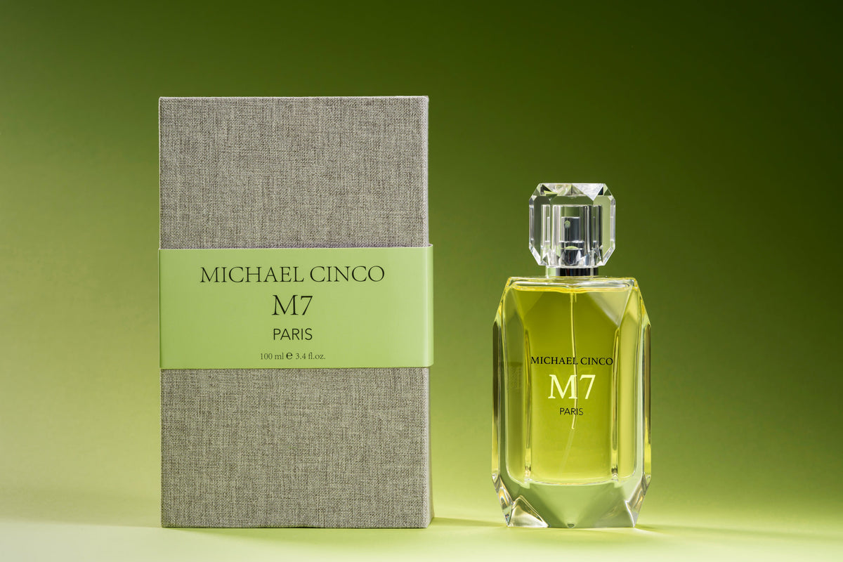 A bottle of M7 Eau de Parfum by Fragrapedia Haus with a faceted glass cap stands beside its textured grey box with a green label, both displayed against a green gradient background.