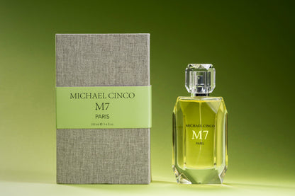A bottle of M7 Eau de Parfum by Fragrapedia Haus with a faceted glass cap stands beside its textured grey box with a green label, both displayed against a green gradient background.