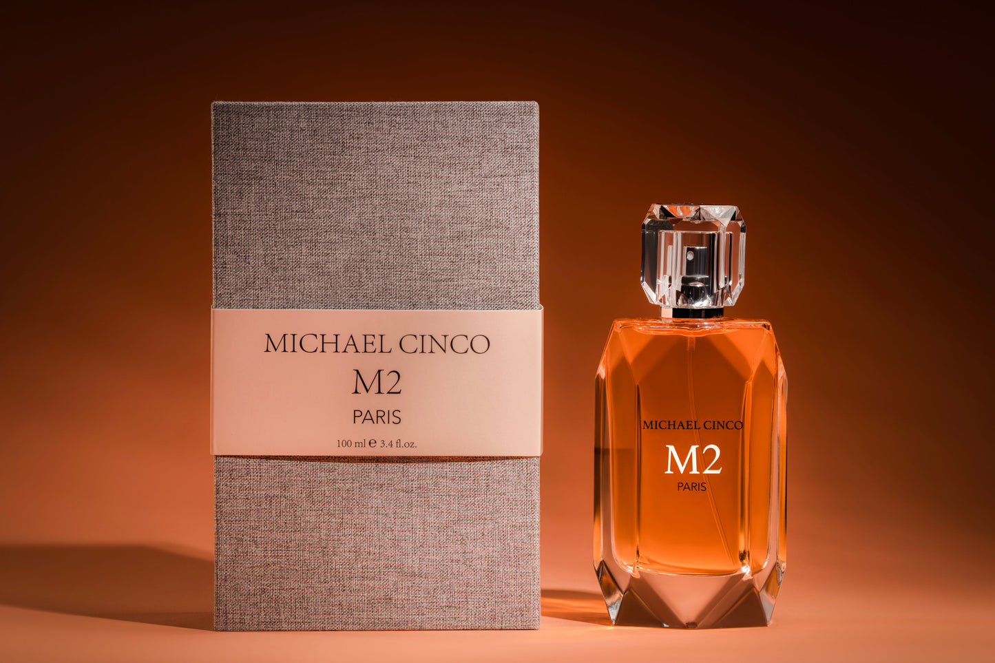 A bottle of Fragrapedia Haus M2 Eau de Parfum by Michael Cinco, filled with amber liquid, stands by its textured gray box on a brown surface, reflecting an exquisite fragrance inspired by oriental spirituality.