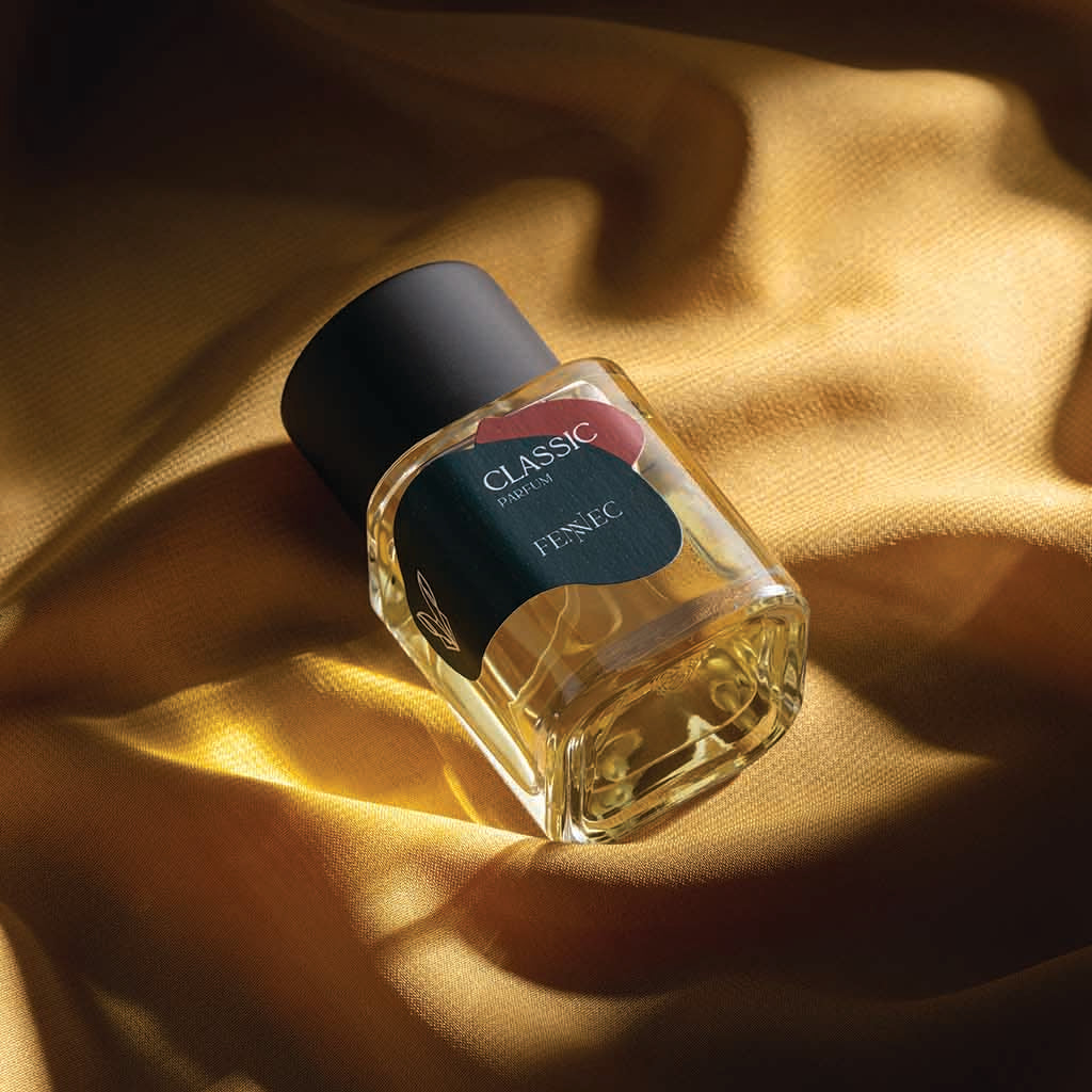 A clear glass bottle of Classic Parfum by Fennec with a black cap rests on gold satin fabric, illuminated by warm light that evokes the elegance of a North African desert fox.