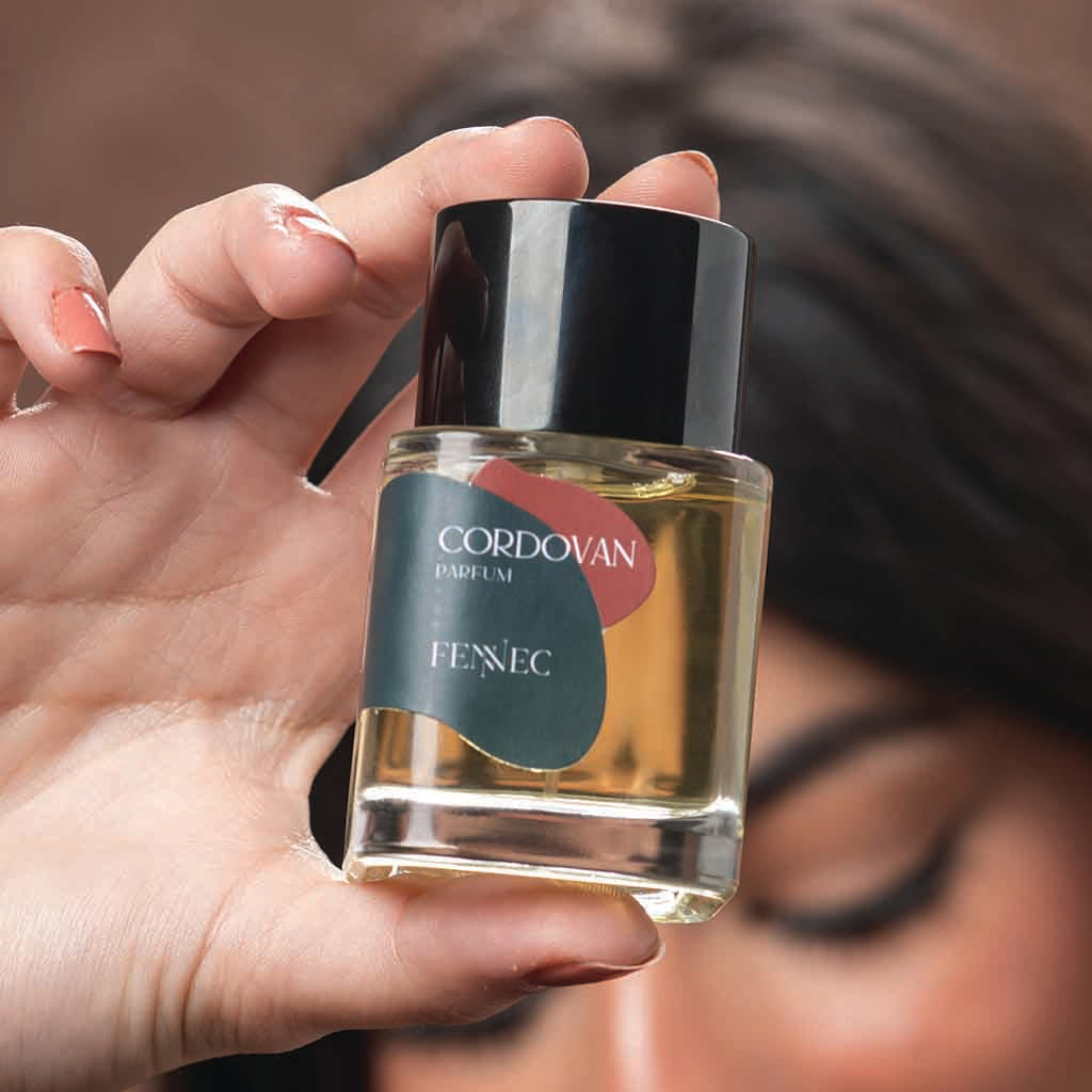 With their face blurred in the background, a person with manicured nails holds up a bottle of Fennec Cordovan Parfum.