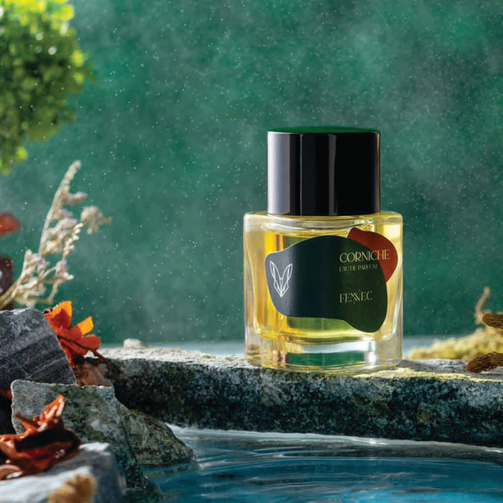 A glass bottle of Fennec’s Corniche EDP rests on a rock near a pool, surrounded by green foliage and flowers. Sunlight filters through, evoking the lush North African landscape that inspired this scent.