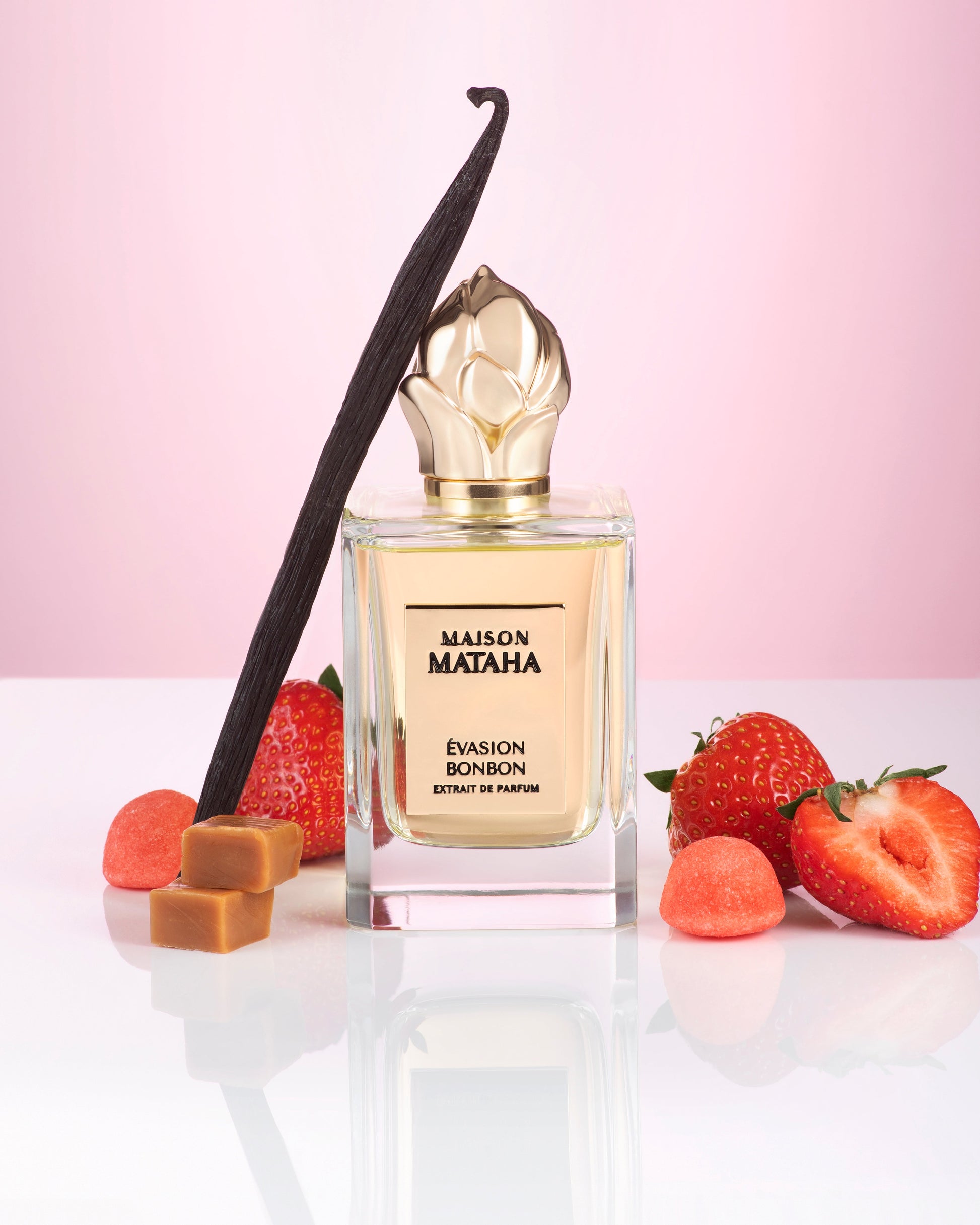 The Evasion Bonbon Extrait de Parfum by Maison Mataha is displayed on a pink background, surrounded by strawberries, caramel cubes, a vanilla bean, and strawberry-shaped candies, evoking a comforting candy-inspired sweetness.