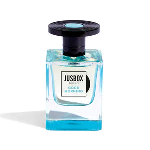 The Jusbox Good Morning Eau de Parfum comes in a clear glass bottle with aqua blue liquid, a round black cap, and a white label displaying the Jusbox brand and fragrance name, evoking fresh magnolia and subtle musk notes.