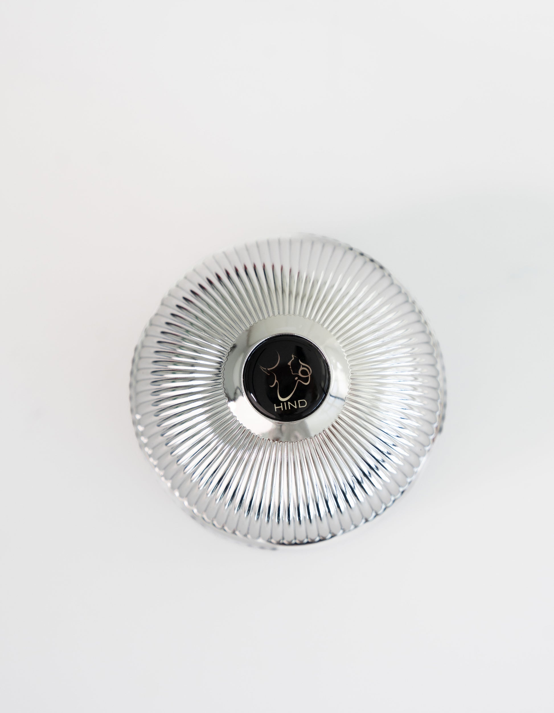 A silver, ribbed, round container with a black center displaying a line drawing of a face and "HIND" on white background, inspired by the luxurious Ahojas Oud Mubakhar from Hind Al Oud.