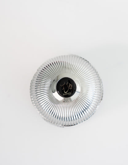 A silver, ribbed, round container with a black center displaying a line drawing of a face and "HIND" on white background, inspired by the luxurious Ahojas Oud Mubakhar from Hind Al Oud.