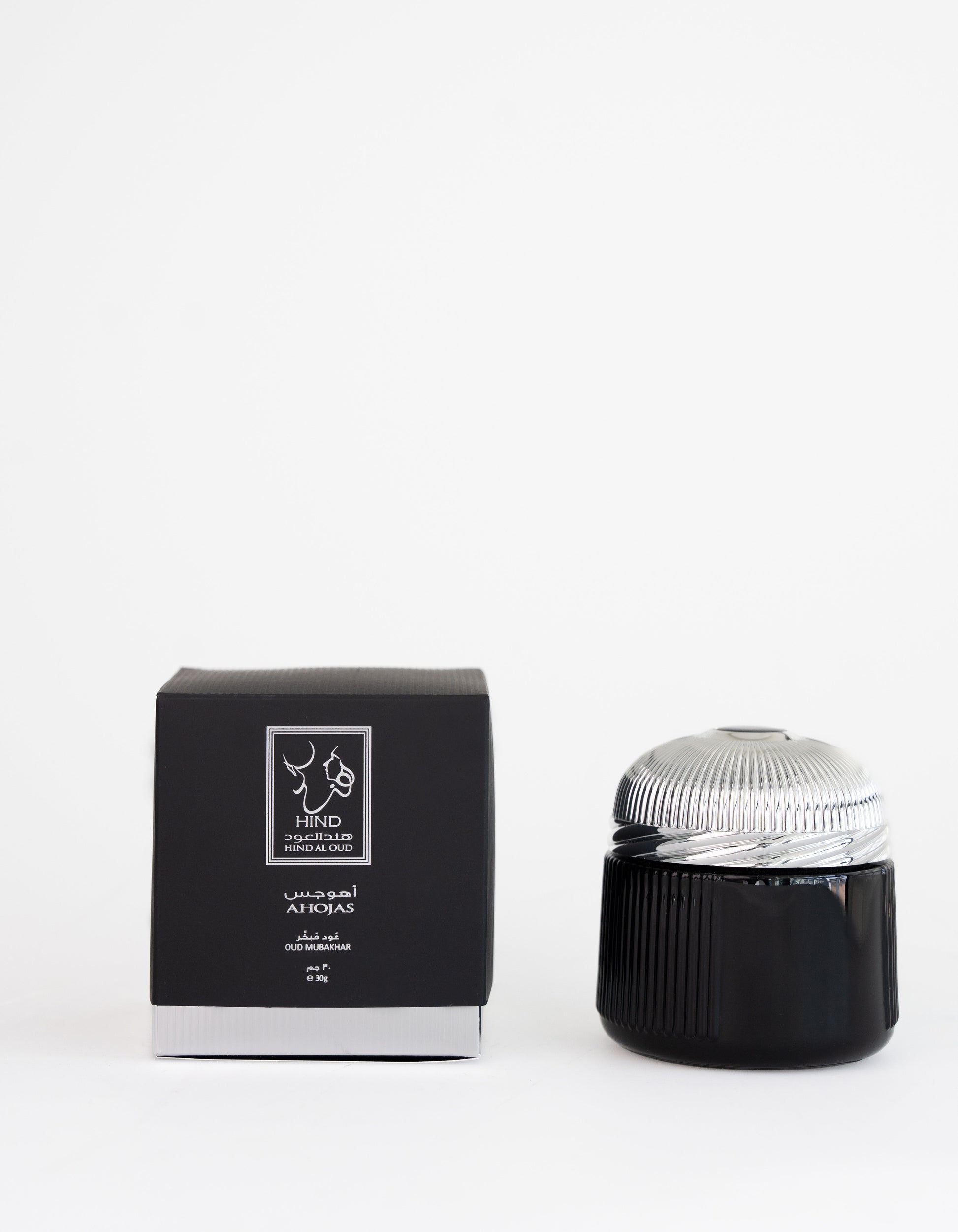 A black and silver Ahojas Oud Mubakhar jar by Hind Al Oud is displayed next to a matching black box with white text and the fragrance logo, set against a plain white background.