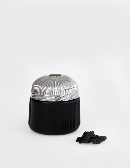 A sleek black and silver cylindrical container with a ridged metal lid sits beside a piece of dark material, capturing the luxurious essence of Ahojas Oud Mubakhar by Hind Al Oud.