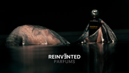 A hand emerges from dark water, holding the ENIGMATIC AURA Extrait de Parfum - Reinvented bottle by REINVĒNTED, with the brand bold at the bottom center—an image evoking luxury and vegan elegance.