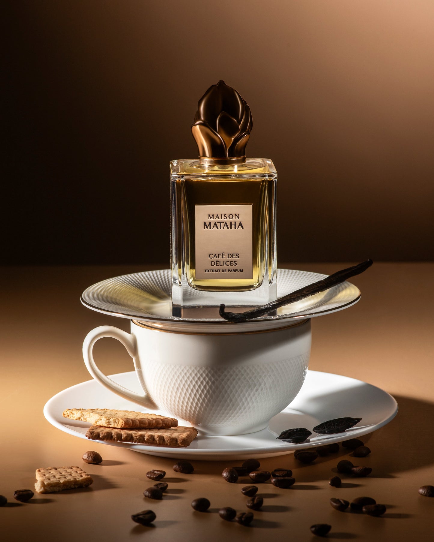 A bottle of Café des Délices Extrait de Parfum by Maison Mataha rests on a plate atop a white teacup, surrounded by coffee beans, cookies, vanilla beans, and chocolate against a warm brown backdrop—evoking rich gourmand allure.