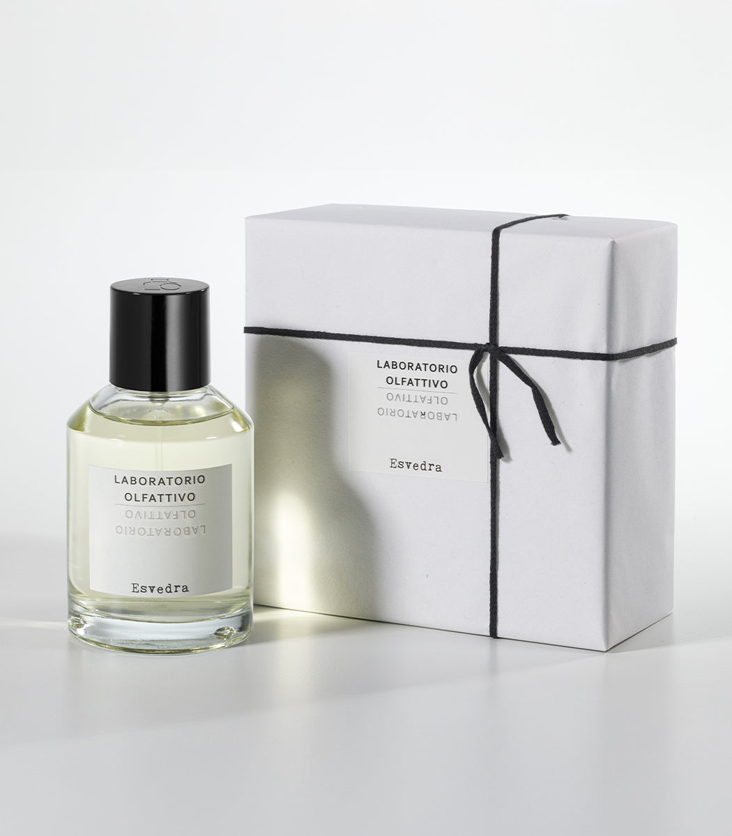 A clear glass bottle of Esvedra by Laboratorio Olfattivo, inspired by the Mediterranean isle Es Vedra with wild vetiver notes, stands next to a neatly wrapped white box tied with black string on a plain white background.