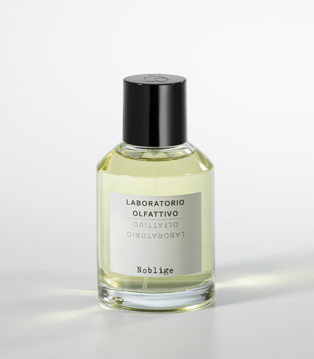 A clear glass bottle of citrus yellow Noblige- Laboratorio Olfattivo perfume, with a black cap and white label, sits on a white surface, evoking the refined aura of an iris lavender fragrance.