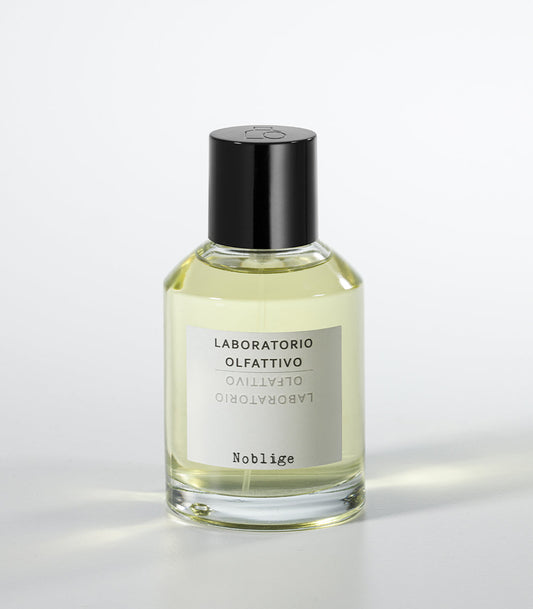 A clear glass bottle of citrus yellow Noblige- Laboratorio Olfattivo perfume, with a black cap and white label, sits on a white surface, evoking the refined aura of an iris lavender fragrance.