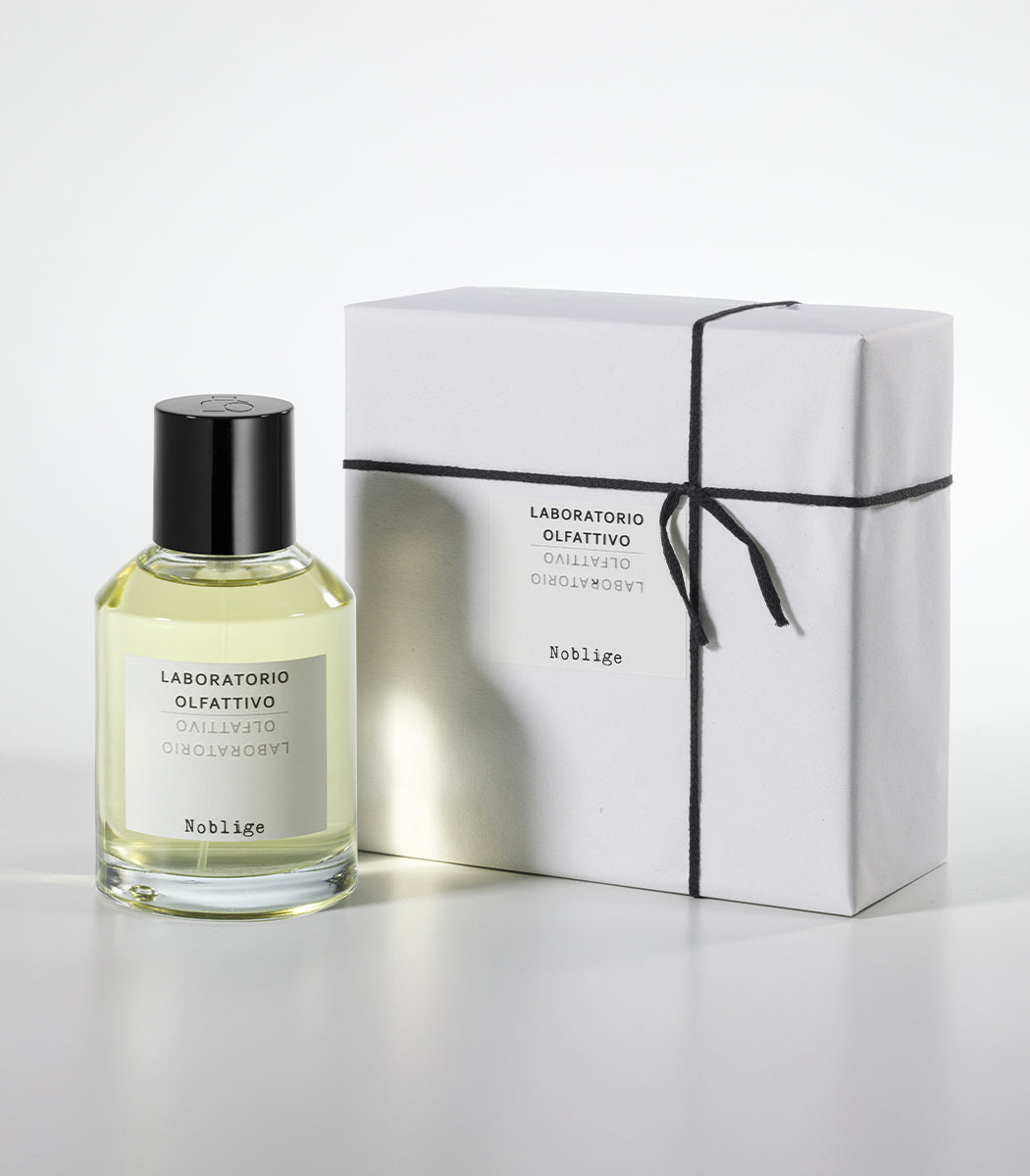 A bottle of Noblige by Laboratorio Olfattivo, an elegant iris and lavender scent, stands beside a white rectangular gift box tied with black string on a plain white background.