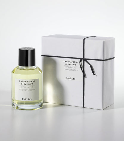 A bottle of Noblige by Laboratorio Olfattivo, an elegant iris and lavender scent, stands beside a white rectangular gift box tied with black string on a plain white background.