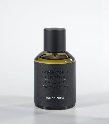 A bottle of Oud in White Eau de Parfum by Fragrapedia Haus, featuring a black cylindrical cap and textured dark gray label against a light background—an elegant floral fragrance with soft sandalwood notes.