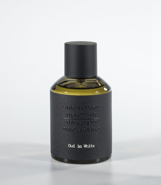 A bottle of Oud in White Eau de Parfum by Fragrapedia Haus, featuring a black cylindrical cap and textured dark gray label against a light background—an elegant floral fragrance with soft sandalwood notes.
