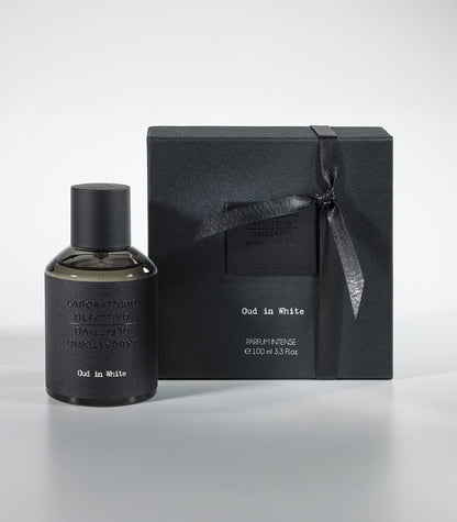 A bottle of Oud in White Eau de Parfum by Fragrapedia Haus sits beside its matching black ribbon-tied gift box, both arranged on a light gray surface.