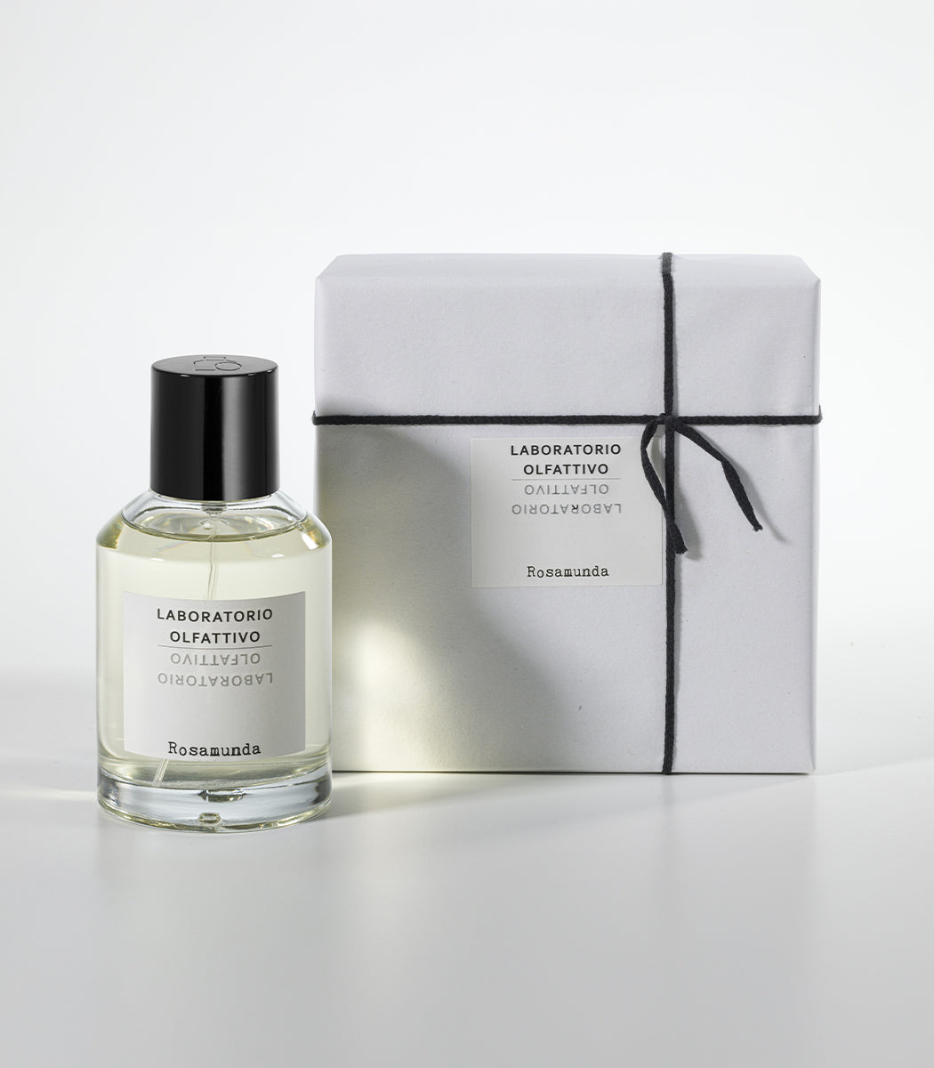 A clear bottle of Rosamunda by Laboratorio Olfattivo with a black cap stands beside a white, black-string-wrapped box, both displayed on a white background to showcase the fragrance’s elegance.