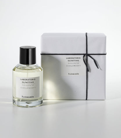A clear bottle of Rosamunda by Laboratorio Olfattivo with a black cap stands beside a white, black-string-wrapped box, both displayed on a white background to showcase the fragrance’s elegance.