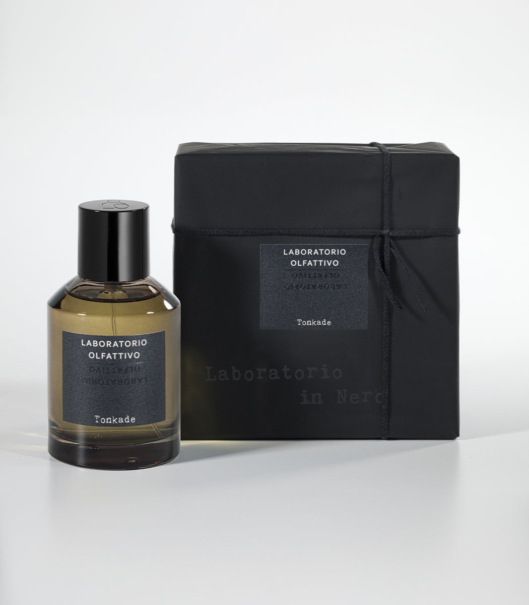 A bottle of Laboratorio Olfattivo Tonkade Eau de Parfum, featuring warm Tonka Bean notes, stands beside its matching black gift-wrapped box with a label, both set on a white surface against a neutral background.