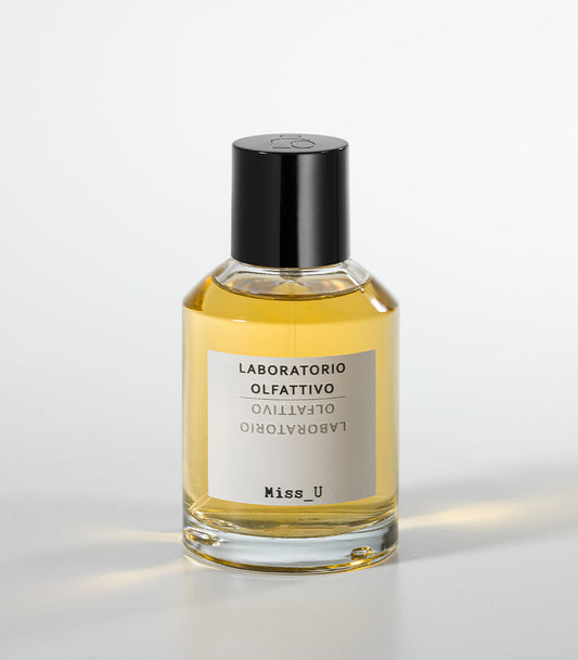 A clear glass bottle of MISS U Eau de Parfum by Laboratorio Olfattivo, filled with yellow liquid and topped with a black cap, stands against a white background—an amber fragrance capturing warm woods and sensual notes.