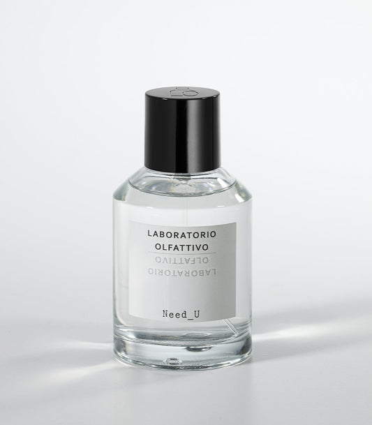 A clear glass bottle of NEED U Eau de Parfum by Laboratorio Olfattivo, with a black cap and labeled branding, rests on a white surface—an elegant unisex fragrance with soft jasmine notes.