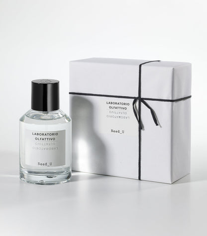 A bottle of Laboratorio Olfattivo NEED U Eau de Parfum stands beside a white, neatly wrapped box tied with black string on a light gray background.