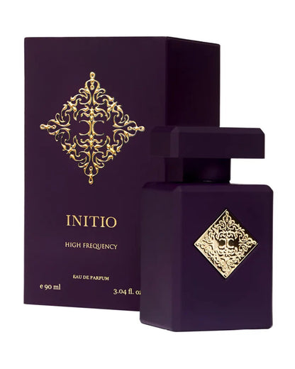 A dark purple perfume bottle with ornate gold accents and a square label stands before a matching box, both reading "High Frequency Eau de Parfum - INITIO," an elegant floral fragrance with magnolia notes.