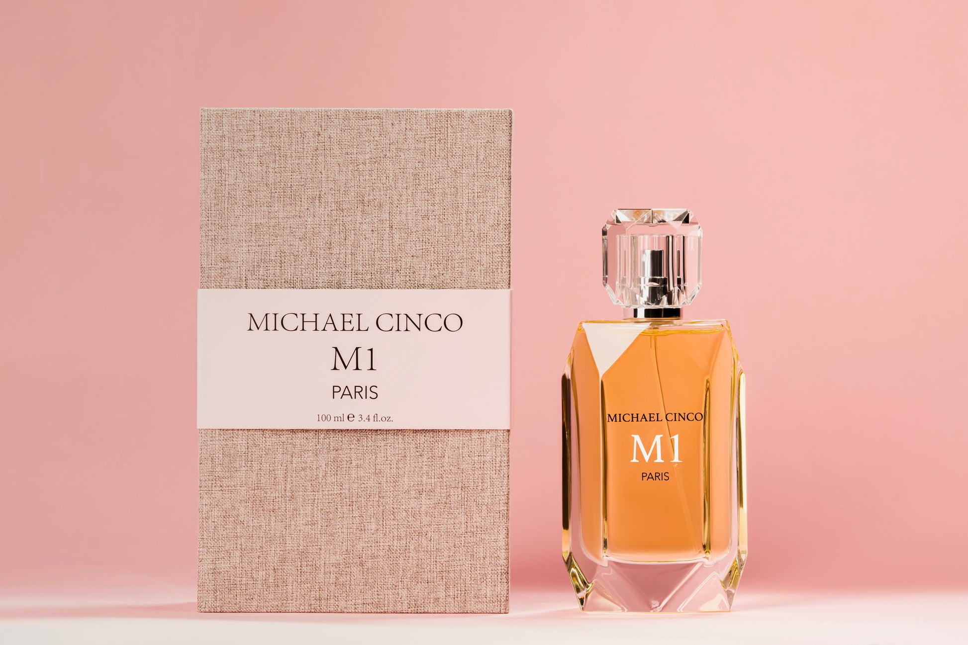 A bottle of M1 Eau de Parfum by Fragrapedia Haus stands beside its textured beige and white box on a soft pink background, showcasing golden liquid and a faceted clear cap, evoking rose and jasmine in a floral fragrance.