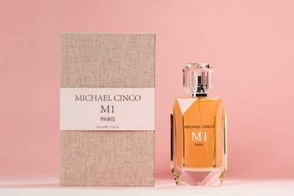 A bottle of M1 Eau de Parfum by Fragrapedia Haus stands beside its textured beige and white box on a soft pink background, showcasing golden liquid and a faceted clear cap, evoking rose and jasmine in a floral fragrance.