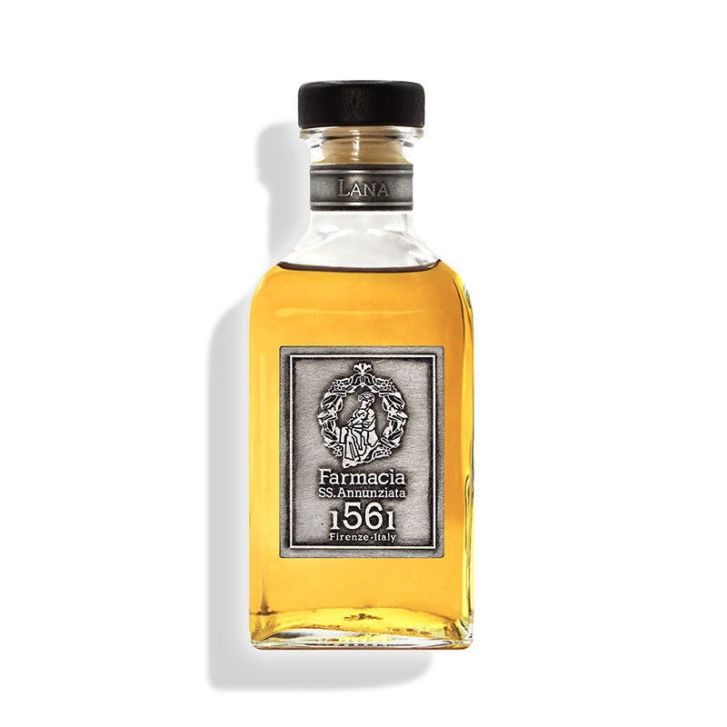 A clear glass Lana DIFFUSER by Farmacia SS. Annunziata 1561, filled with golden liquid and topped with a black cap, features a silver label. The bottle casts a soft shadow on white, exuding refined Arte della Lana elegance.