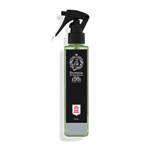 A green transparent silk Seta Room Spray bottle by Farmacia SS. Annunziata 1561 has a black trigger cap and black label with a white ornate logo, the brand’s name, and a small red and white emblem near the bottom.
