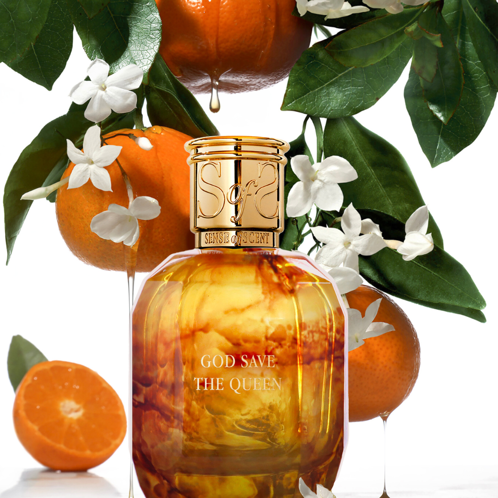A golden GOD SAVE THE QUEEN Eau de Parfum by Sense of Scent stands among whole and sliced oranges, green leaves, and white blossoms, while droplets of orange juice fall above and around the bottle.
