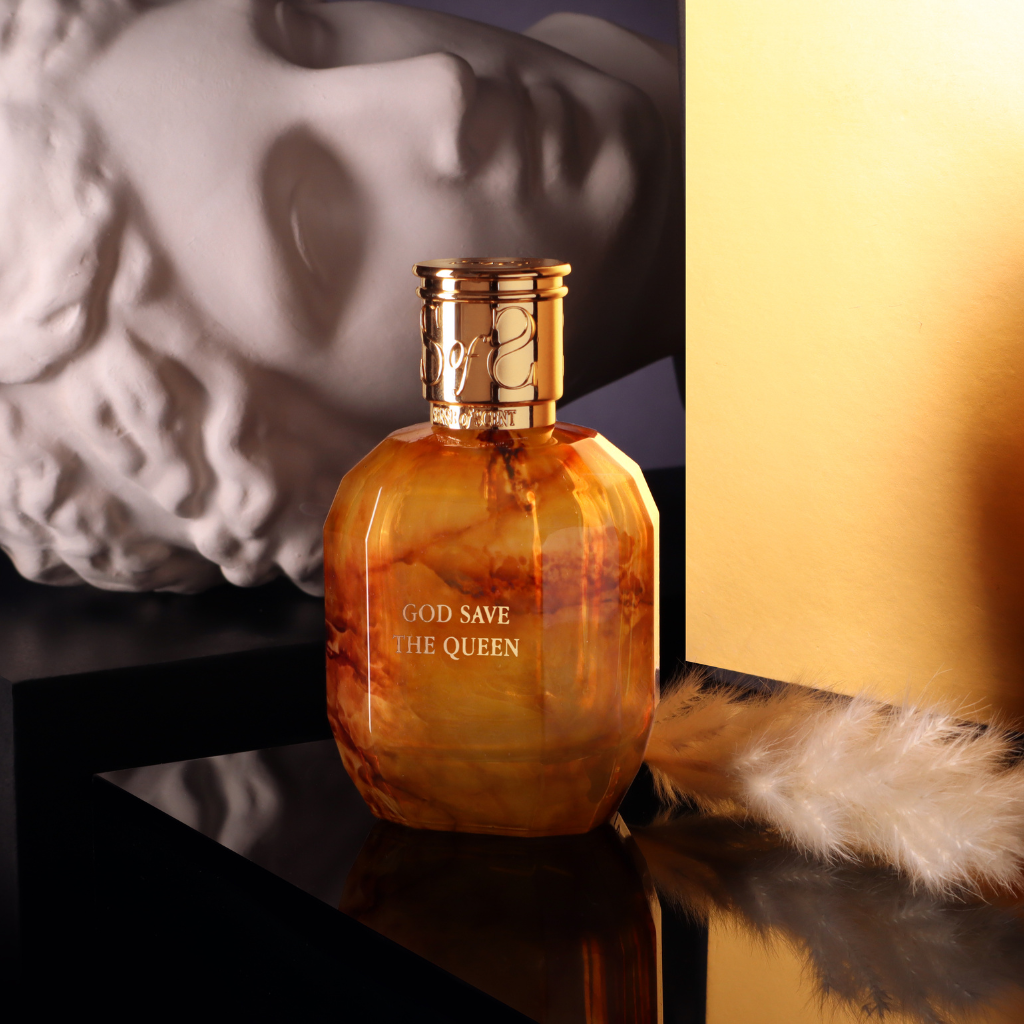 A golden-brown GOD SAVE THE QUEEN Eau de Parfum by Sense of Scent sits on a glossy black surface next to a white feather, with a white classical statue head and yellow panel in the background.