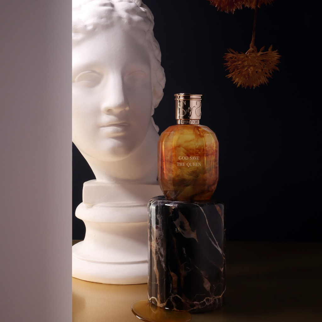A bottle of GOD SAVE THE QUEEN Eau de Parfum by Sense of Scent rests on a black marble pedestal next to a white classical bust, with dried flowers above and a dark backdrop.