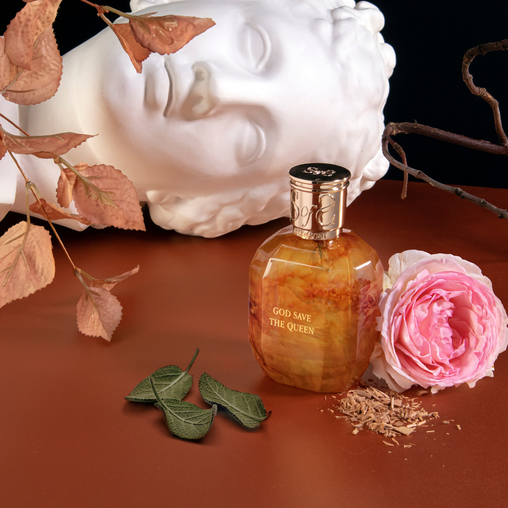 A marble bust lies behind a brown Sense of Scent GOD SAVE THE QUEEN Eau de Parfum bottle. Beside it sit a pink rose, dried leaves, twigs, and petals on a brown surface against a dark background.