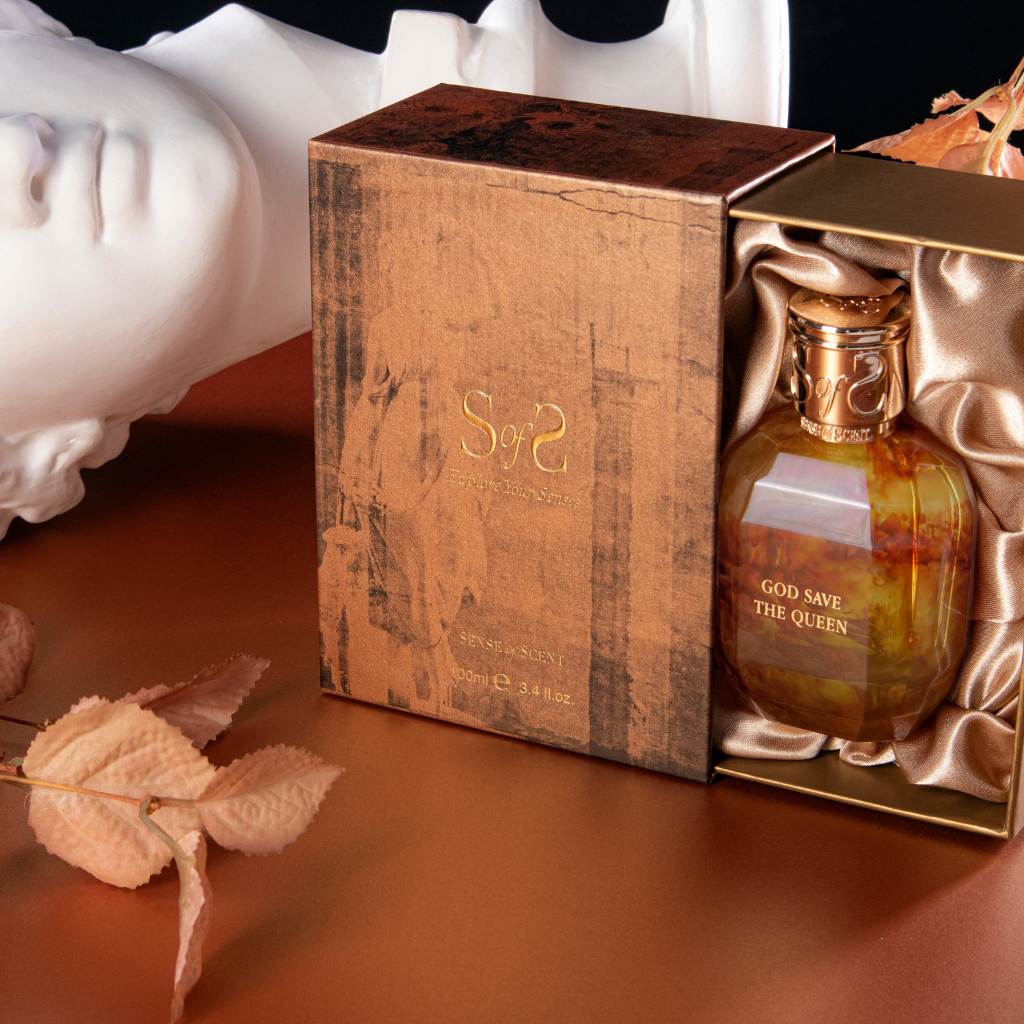 A gold and brown GOD SAVE THE QUEEN Eau de Parfum box by Sense of Scent sits open on a metallic surface, displaying its glass bottle in gold satin. Dried leaves and a white classical statue are visible in the background.