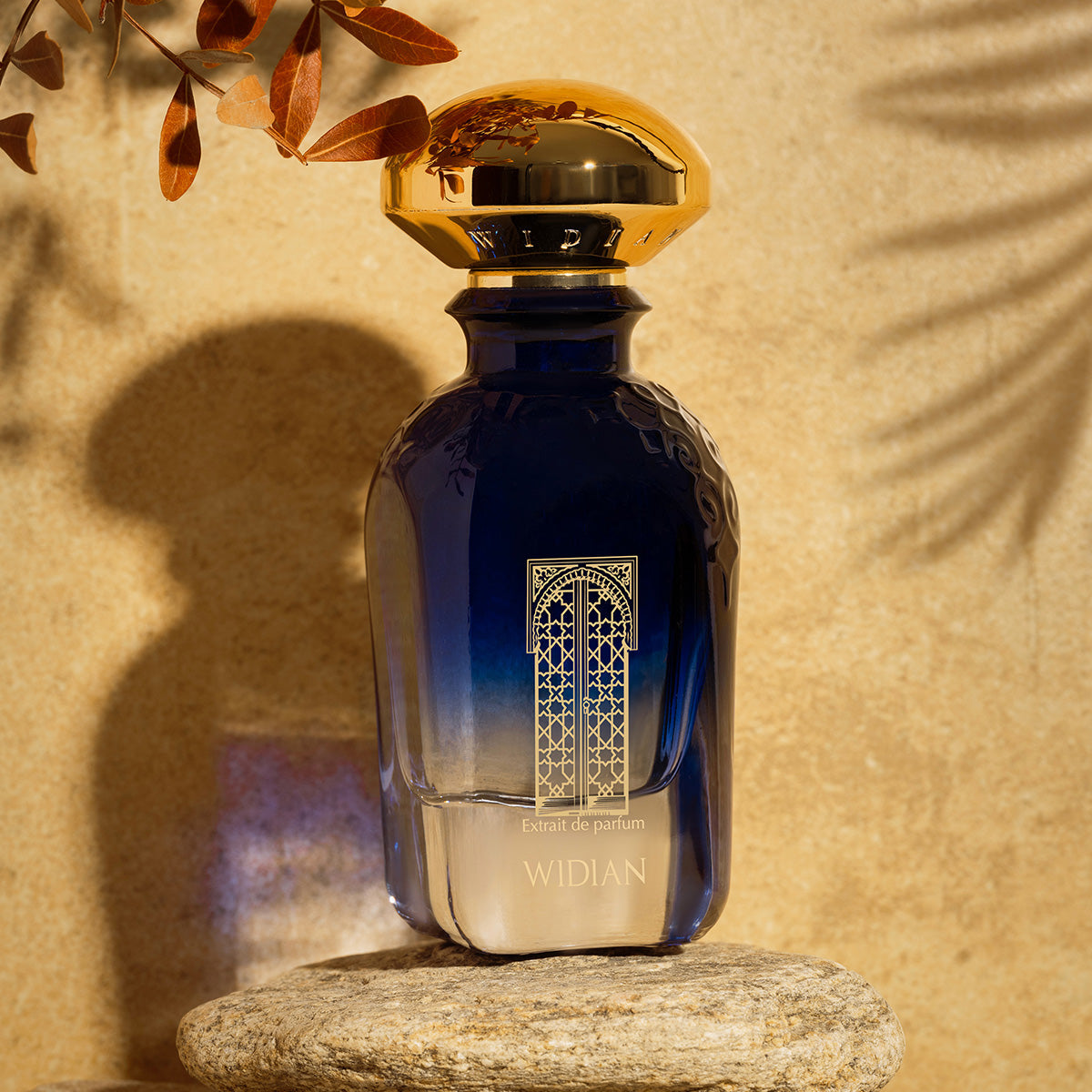 A luxurious blue Granada Extrait de Parfum bottle by Fragrapedia Haus with a gold cap stands on stone amid sunlight and brown leaves, evoking soft notes of rose and juniper for an elegant, inviting atmosphere.