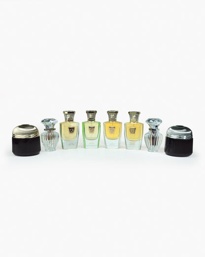 Seven elegant bottles from the Hind Al Oud Immortal Allure Collection Gift Set stand in a row against a white background, containing gold and green perfumes. Two bottles have ribbed designs, each topped with a decorative cap.