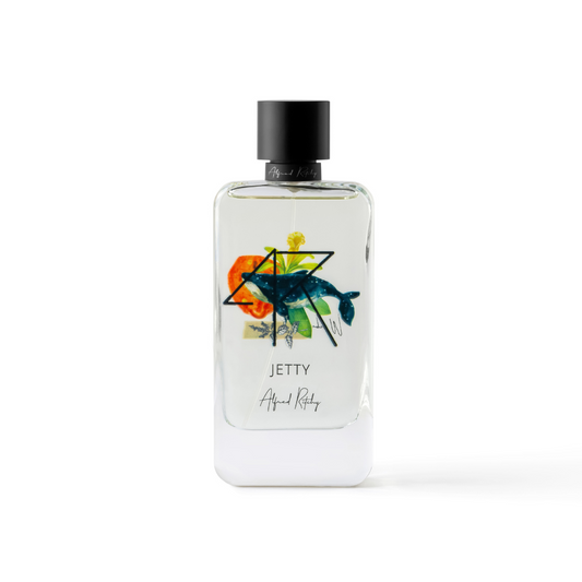 A clear glass JETTY Eau de Parfum by Alfred Ritchy features colorful abstract art with a fish, geometric shapes, and a black cap—reflecting fresh bergamot and smooth sandalwood notes.
