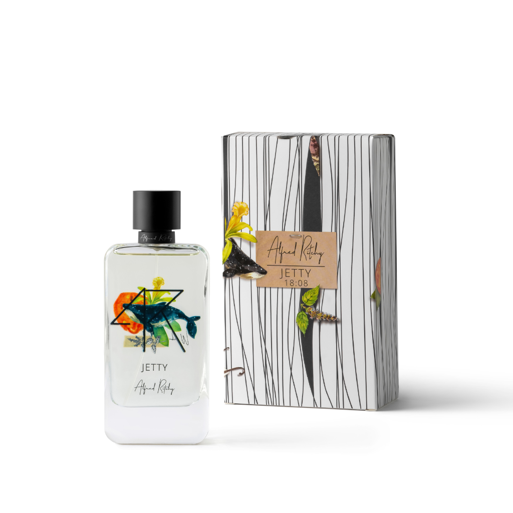 A bottle of Alfred Ritchy's JETTY Eau de Parfum, featuring a vibrant fish illustration, stands beside its white box with black stripes and a brown label. Floral accents and sandalwood notes complete this distinctive fragrance presentation.