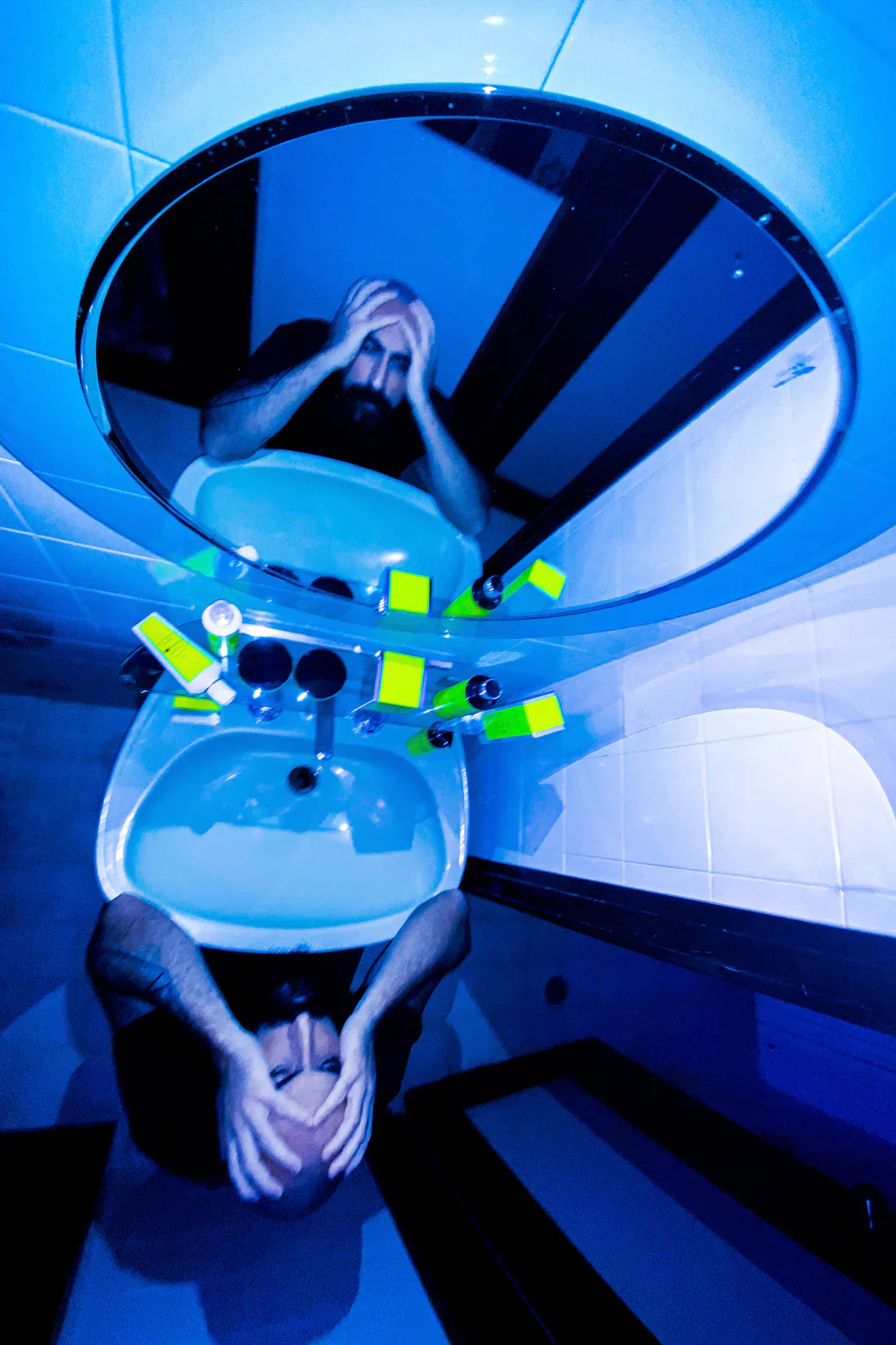 A person stands over a bathroom sink, head in hands, reflected in a circular mirror. Blue light bathes the scene, with yellow items—including a bottle of Filippo Sorcinelli SUPERFLUO LAVS SHAMPOO—set around the sink.
