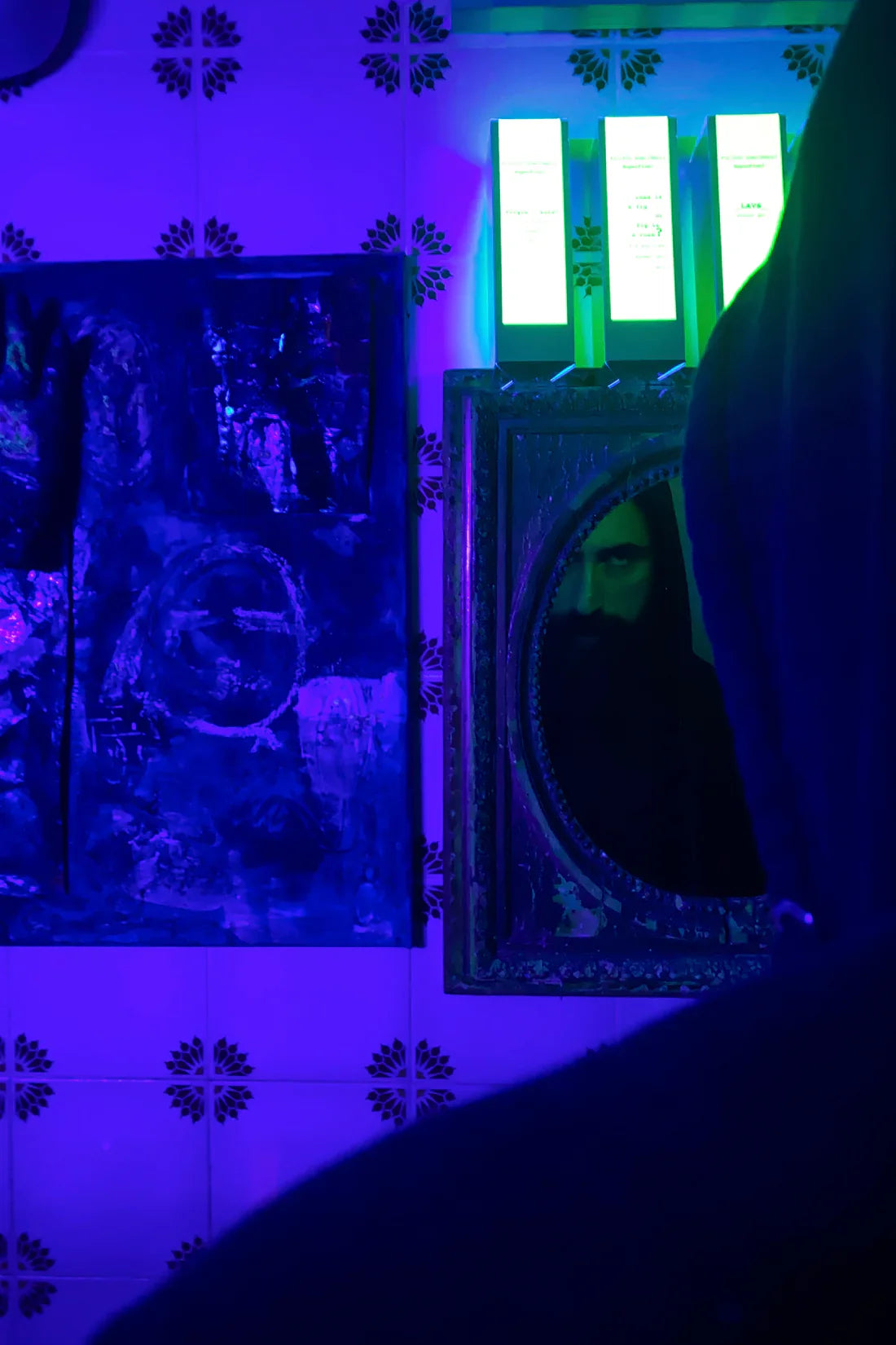 A hooded figure is reflected in a small ornate mirror on tiled walls lit by blue and purple light. Green neon, abstract art, and incense set the mood of Filippo Sorcinelli’s SUPERFLUO LAVS SHOWER GEL experience.