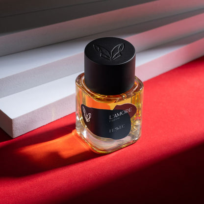 A glass bottle of Fennec L'Amour EDP with a black cap rests on a red surface near white slatted blinds, sunlight casting shadows—its scent evoking the mystique of the North African desert.