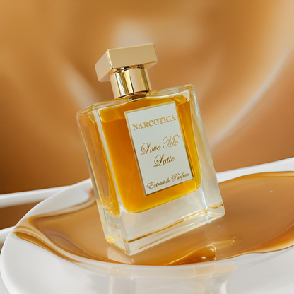 A bottle of Narcotica Love Me Latte Eau de Parfum rests on a white surface with golden liquid pooling around it, set against a warm, blurred brown background.