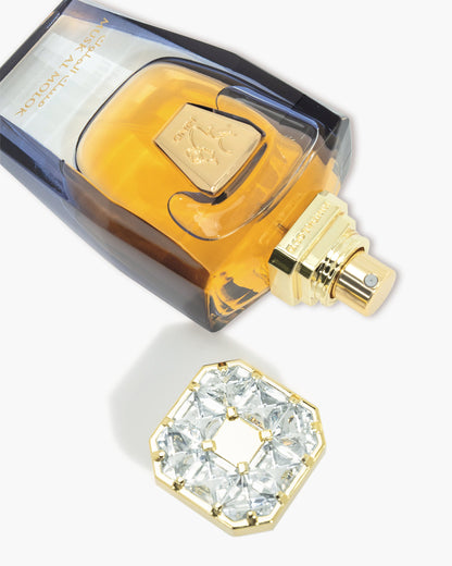 A luxurious bottle of Musk Al Molok Parfum by Hind Al Oud, with gold accents and amber liquid, rests beside its ornate gold and crystal hexagonal cap on a white background—exuding elegance and sophistication.
